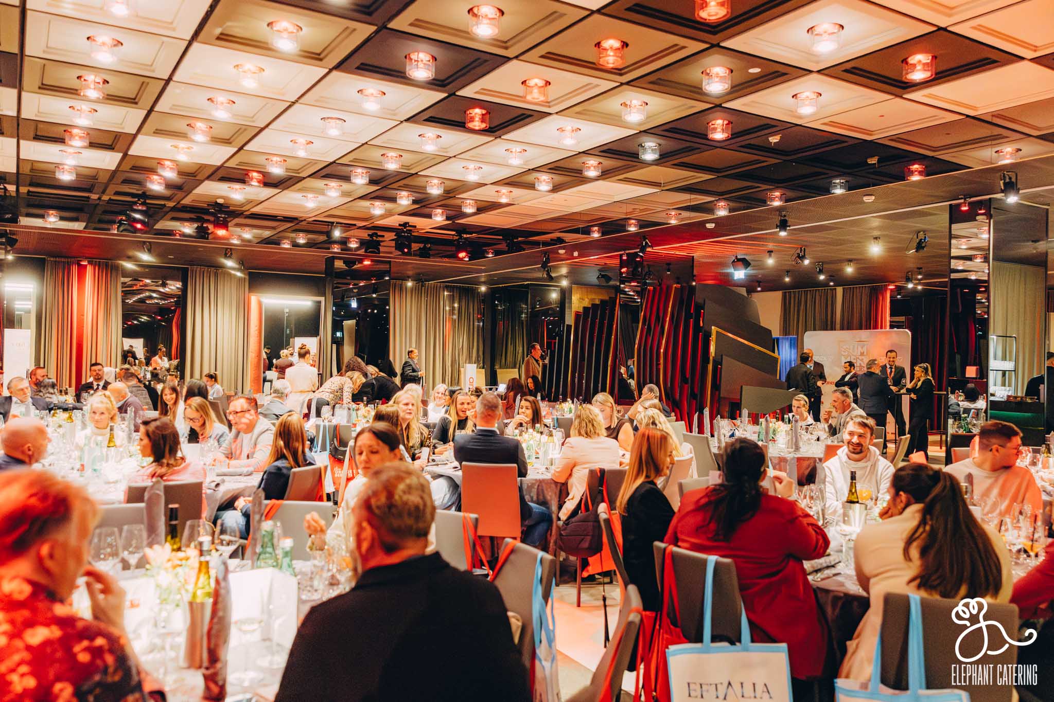 Banquet hall full of round tables with guests dining under warm lighting, Elephant Catering branding visible in the corner.