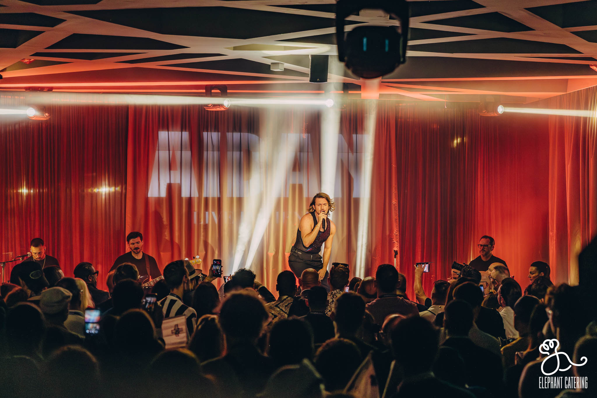 Singer center stage performing to a crowded audience, red curtains behind, bright stage lights streaking across the scene.