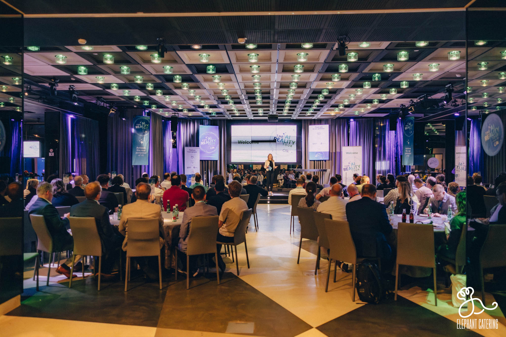 Large conference hall filled with seated attendees facing a stage, a speaker at the podium beneath a big'Welcome' screen and banners reading 'The AI Reality Tour'
