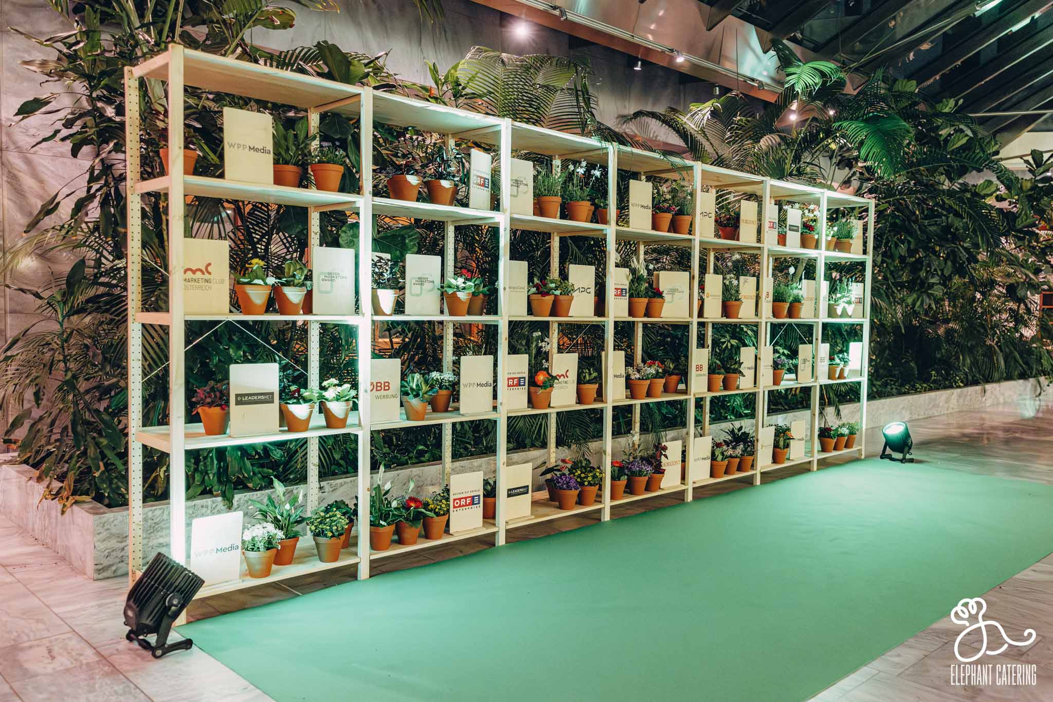 Indoor plant display on white metal shelves with numerous small potted plants and sponsor signs, set on a green carpeted runway at an event.
