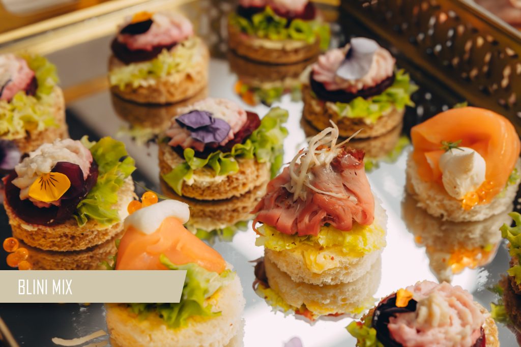 Assorted blini bites with smoked salmon, greens, and creamy toppings on a mirrored tray labeled BLINI MIX.