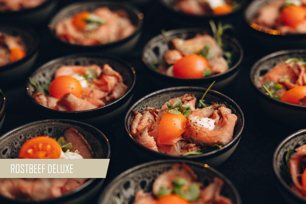 Small black bowls filled with roast beef, cherry tomatoes, greens, and a dollop of creamy topping on a dark surface.