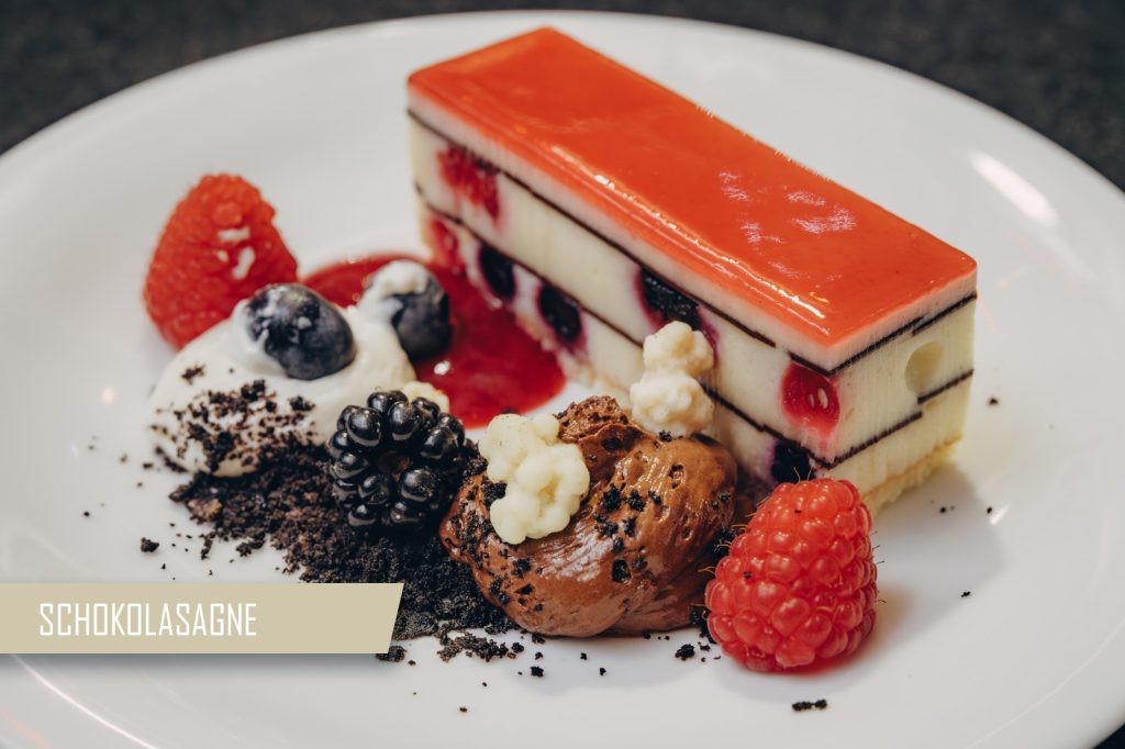 Rectangular berry cheesecake with orange glaze, layered with fruit, on a white plate, surrounded by berries and crumbs.